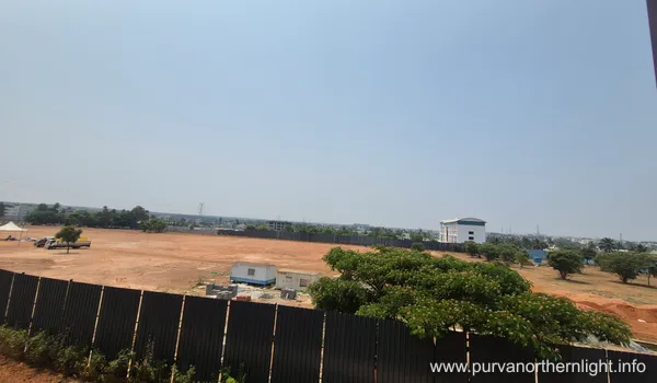 A wide-angle, elevated view of the expansive Purva Northern Lights project site under a clear blue sky. The ground is leveled with reddish-brown soil, featuring a temporary white site office, construction fencing in the foreground, and lush green trees bordering the perimeter.