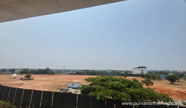 A panoramic daytime view of the Purva Northern Lights development land. The image shows a cleared construction area secured by dark metal fencing, with scattered greenery and a white multi-story building visible in the distant background under a bright, hazy sky.